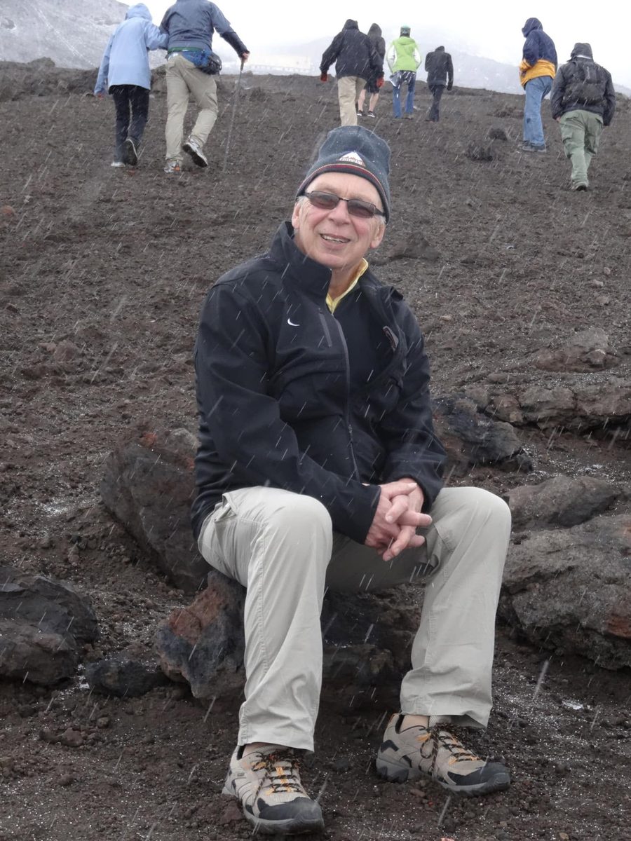 Jim Jensen sitting on volcanic rock in falling sleet, smiling, with hikers climbing the slope behind him